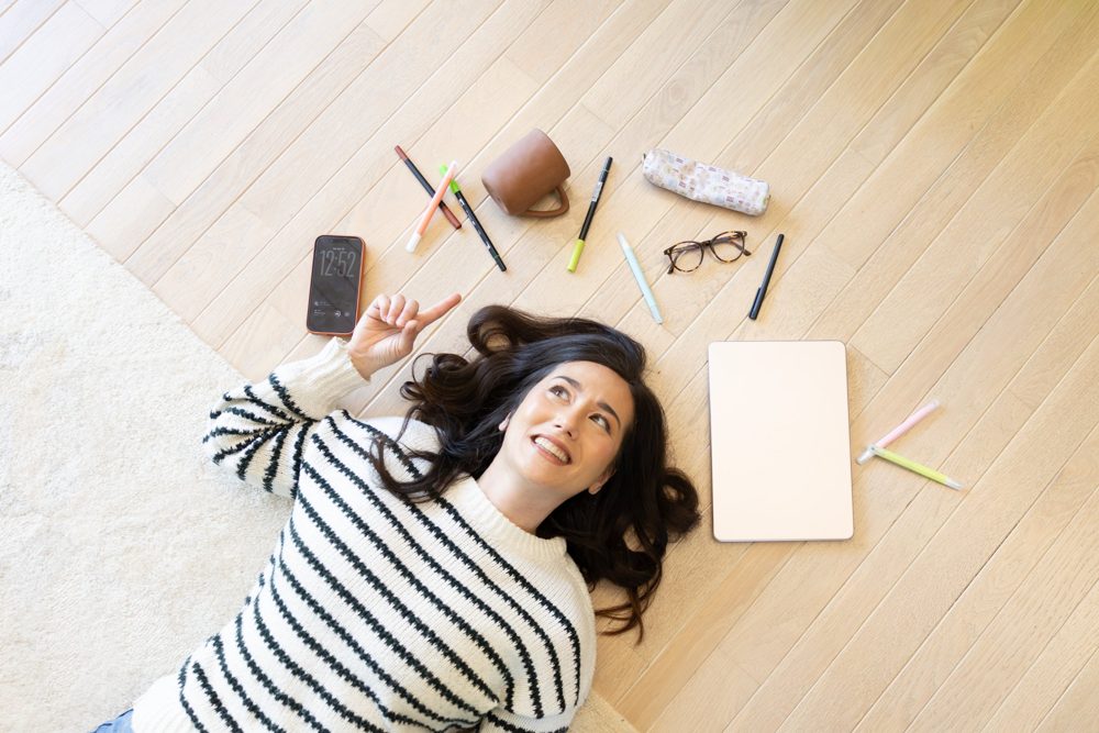 Sondra lying on the floor pointing up at a ring of pens, a phone, glasses and a closed laptop
